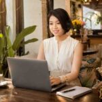 How to Make Your Laptop Faster for Daily Work Woman works on a laptop with headphones in a sunny café.